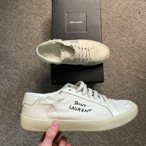 Vita sneakers från Saint Laurent - Snygga vita sneakers från Saint Laurent med en sliten look och handskriven logga på sidan. Skorna har snörning och är tillverkade i ett slitstarkt material. Perfekta för en avslappnad stil.