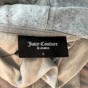 Grå hoodie från Juicy Couture - Säljer en snygg grå hoodie från Juicy Couture med dragkedja och huva som jag köpt här på plick men enbart testat då den var för liten för mig. Tröjan har en stilren design med ett litet broderat emblem på bröstet och en dekorativ dragkedja med ett 'J'-format hänge. Perfekt för en avslappnad stil. Ställ gärna frågor om det är nått ni undrar!