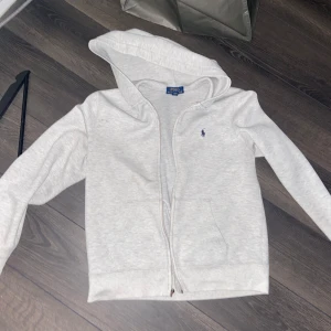 Grå hoodie från Polo Ralph Lauren - Drag kedjan funkar men den där zip grejen som man drar upp o nere me är borta. Säljer en stilren grå hoodie från Polo Ralph Lauren med dragkedja och huva. Tröjan har långa ärmar och en liten broderad logga på bröstet. Perfekt för en avslappnad look. ANDRA GÅNGEN GILLIGT!