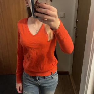 Orange tröja från Peak Performance - Säljer en snygg orange/röd tröja från Peak Performance i storlek S. Tröjan har en v-ringad design och långa ärmar, perfekt för en stilren look. Den är tillverkad i ett mjukt material och har en liten broderad logga på framsidan. Passar perfekt till jeans eller kjol!