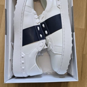 Valentino Garavani sneakers med blå detalj - Snygga vita sneakers från Valentino Garavani med en bred blå rem över ovansidan. Skorna har en stilren design med nitar på sidorna och en robust sula. Perfekta för en trendig look. Ej använda samt helt nya. Pris kan diskuteras vid snabb affär!