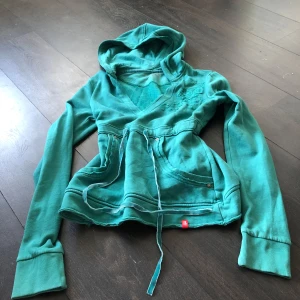 Turkos hoodie med broderi från EDC - Säljer en snygg turkos hoodie från EDC med broderade detaljer på bröstet. Hoodien har en v-ringad design och dragsko i midjan för en justerbar passform. Kan färga av sig på kroppen! 