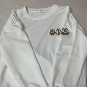 Vit långärmad tröja från Moncler - Säljer en stilren vit långärmad tröja från Moncler med broderad logga på bröstet. Perfekt för en avslappnad och trendig look. Tröjan har en rund halsringning och är tillverkad i ett mjukt material.