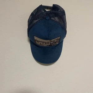 Blå keps från Emilio Pendenza  - Snygg blå keps från Emilio Pendenza med ett unikt tryck av en strandscen på framsidan. Kepsen har ett nätmaterial på baksidan för bättre ventilation. Perfekt för en avslappnad stil.