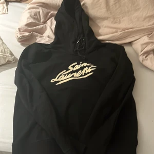 Svart hoodie från Saint Laurent - Snygg svart hoodie från Saint Laurent med deras ikoniska logga i vitt på bröstet. Perfekt för en stilren och avslappnad look. Den har en klassisk passform med långa ärmar och en praktisk huva med snörning.