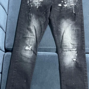 Svarta slitna jeans med raka ben - Säljer ett par svarta jeans med slitningar och små färgstänk framtill. Jeansen har raka ben, klassisk femficksmodell och knappgylf. Perfekta för en cool och avslappnad stil.