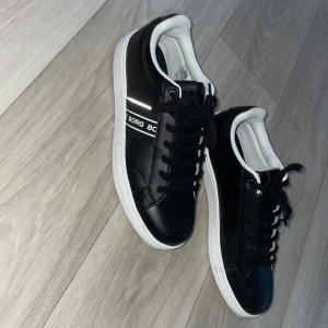 Svarta sneakers från Björn Borg - Snygga svarta sneakers från Björn Borg med vita detaljer och vit sula. Skorna har snörning och en klassisk rund tå. Perfekta för en stilren och sportig look. Märkeslogga syns på sidan och innersulan.