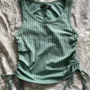 Grön ribbad linnetopp från SHEIN - Säljer en ljusgrön ribbad linnetopp från SHEIN med knytband i sidorna. Toppen är ärmlös och har en rund halsringning. Perfekt för varma dagar och enkel att matcha med olika outfits.