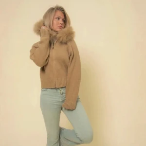 Alessa furjacka - Säljer en beige stickad jacka med dragkedja och stor krage med fuskpäls. Jackan är croppad och har långa ärmar med breda muddar. Perfekt för kyligare dagar och ger en trendig look.