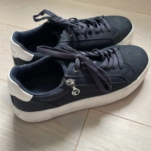Mörkblåa sneakers från Tamaris med vita detaljer - Säljer ett par mörkblåa sneakers från Tamaris med vita detaljer på hälen och vit sula. Skorna har snörning, dragkedja på sidan och en liten metallcharm. Perfekta för en stilren vardagslook.
