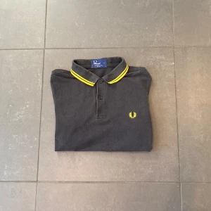 Svart pikétröja från Fred Perry - Säljer en svart pikétröja från Fred Perry i slim fit med klassisk krage och guldiga/gula ränder på krage och ärmslut. Tröjan har korta ärmar och är tillverkad i 100% bomull. Ikonisk Fred Perry-logga broderad på bröstet. Perfekt till sommaren☀️💯