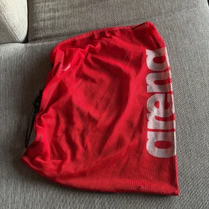 Röd mesh bag från Arena - Säljer en röd mesh bag från Arena med vitt tryck på sidan. Väskan har snörstängning med svarta snören och är tillverkad i ett lätt och luftigt syntetmaterial. Perfekt att bära tränings utrustning eller badkläder i.