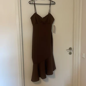 Brun volangklänning från Bubbleroom - Säljer en brun klänning från Bubbleroom med smala axelband och volang nertill. Klänningen har en figurnära passform och asymmetrisk nederkant som ger en snygg siluett. Helt oanvänd. Har storleken S.