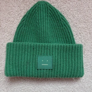 Grön ribbad mössa från Acne Studios - Grön stickad mössa från Acne Studios med ribbad struktur och uppvikt kant. På framsidan sitter en tygpatch med det klassiska ansiktet. Perfekt för att hålla värmen och ge en cool touch till din outfit.