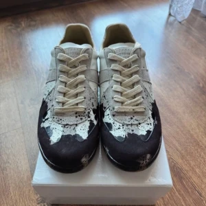 Maison Margiela sneakers med svart och vit färg - Unika sneakers från Maison Margiela i storlek 43. Skorna har en beige bas med svart och vit färg som ser ut som färgstänk. De har snörning, rund tå och en gummisula. Tillverkade i Italien och perfekta för dig som vill sticka ut.