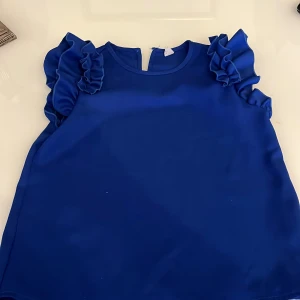 Blå volangtopp - Säljer en snygg blå topp med volangdetaljer på axlarna. Toppen har en rund halsringning och är ärmlös, perfekt för att ge outfiten lite extra edge. vet tyvärr inte märket. Säljer för ”100” kr med frakt. endast använd en gång. Storlek Xs.