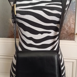 Svart axelväska från Balpri - Säljer en stilren svart axelväska från Balpri, tillverkad i skinn. Väskan har ett klassiskt fyrkantigt format med lock och justerbar axelrem med gulddetalj. Insidan är fodrad i brunt tyg och har en dragkedja.