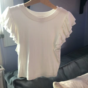 Vit ribbad topp med volangärmar från SHEIN - Säljer en söt vit ribbad topp från SHEIN med volangdetaljer på ärmarna. Toppen har rund halsringning och är tillverkad i ett mjukt, stretchigt material. Perfekt för att skapa en feminin och trendig look.