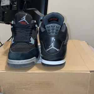 Svarta Nike Air Jordans 4 Retro - Säljer ett par svarta Nike Air Jordans 4 Retro med grå och vita detaljer. Skorna har klassisk mesh och skinn, synlig Air-sula och Jumpman-logga på hälen. Perfekta för dig som gillar streetwear och sneakers med ikonisk design. Inte trasiga nån stans och knappt använda 40-40,5