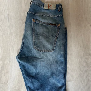 Blå jeans från Nudie Jeans - Säljer ett par klassiska blå jeans från Nudie Jeans med raka ben och normal passform. Jeansen har fem fickor, orangea sömmar och en tydlig Nudie-logga på bakfickan. Perfekta för dig som gillar en tidlös denimlook. Dessa är smått åt det baggiare hållet. Mindre skavanker och fläckar med fortfarande extremt snygga 🔥