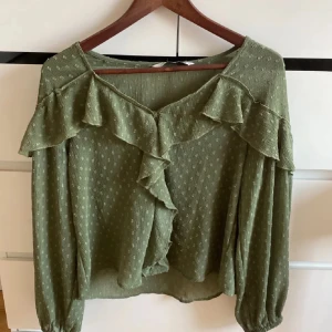 Grön blus med volang från Zara - Säljer en ljusgrön blus från Zara med volangdetaljer längs ringningen och över ryggen. Blusen har långa ärmar med resår vid ärmslut och små prickar i tyget. Medföljer matchande snören med tofsar och metallknappar. Jag har klippt av snörerna men dem är lätta att sy fast!