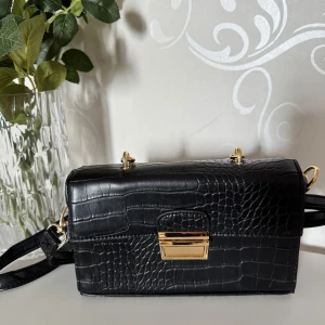Svart axelväska  - Stilren svart axelväska med krokodilpräglat mönster och guldfärgat handtag samt spänne. Väskan har både axelrem och handtag, vilket gör den enkel att bära 🖤