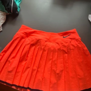 Neonorange plisserad tenniskjol från Nike - Säljer en neonorange tenniskjol från Nike med plisserad design och inbyggda shorts. Kjolen har en svart Nike-logga framtill och är perfekt för dig som vill sticka ut på tennisbanan. Bekväm midja och sportig look.