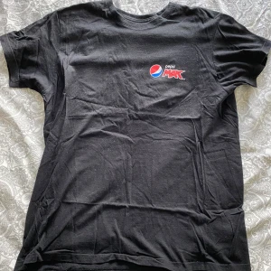 Svart Pepsi Max t-shirt - Svart t-shirt med Pepsi Max-logga tryckt på bröstet. Klassisk passform med rund hals och korta ärmar. Perfekt för dig som gillar snygga och enkla statement-plagg. Tillverkad i mjuk bomull.