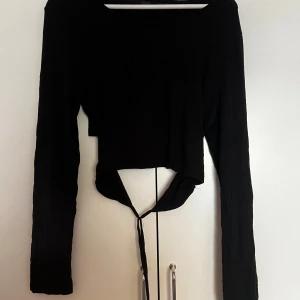 Svart croppad topp med knyt - Säljer en svart långärmad croppad topp med knyt baktill. Toppen har en enkel design med rund halsringning och långa ärmar, perfekt att styla till olika outfits.