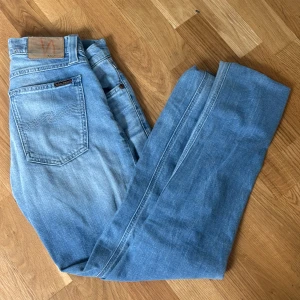 Ljusblå jeans från Nudie Jeans - Säljer ett par ljusblå jeans från Nudie Jeans med klassisk femficksdesign och normal passform. Jeansen har dragkedjegylf och subtila slitningar för en avslappnad look. Perfekta för dig som gillar stilrena och bekväma jeans.