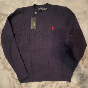 Mörkblå stickad tröja från Polo Ralph Lauren - Säljer en snygg mörkblå stickad tröja från Polo Ralph Lauren med det klassiska röda logotypbroderiet på bröstet. Tröjan har en rund hals och långa ärmar, perfekt för kyligare dagar. Ett stilrent val för garderoben!