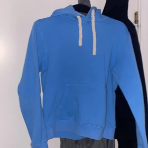 Blå hoodie från Syna Wrld  - Säljer en blå hoodie från SYNA wrld med vit graffiti-tryck på huvan och grova vita snören. Tröjan har känguruficka framtill och är perfekt för en avslappnad stil.