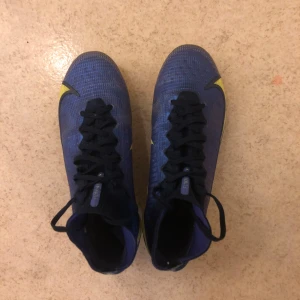 Nike Mercurial Super Fly 8 Elite - Säljer ett par blå Nike Mercurial fotbollsskor med gula detaljer och svart snörning. Skorna har högre strumpkrage för extra stöd och är designade för spel på gräs. Snygg och sportig look med tydlig Mercurial-logga på hälen.