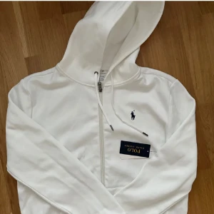 Vit hoodie från Polo Ralph Lauren - Säljer en klassisk vit hoodie från Polo Ralph Lauren med dragkedja och broderad logga på bröstet. Tröjan har långa ärmar och justerbar huva. Perfekt för en stilren och avslappnad look.