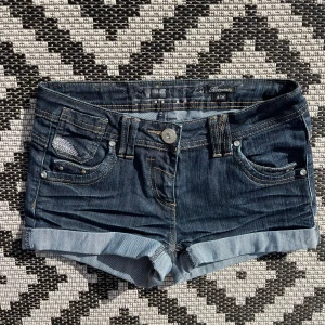 Mörkblå jeansshorts med nitar - Snygga mörkblå jeansshorts med upprullade benslut och dekorativa nitar på bakfickan. Klassisk femficksmodell med ljusa sömmar och knappgylf. Perfekta för sommaren och passar till det mesta.