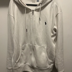 Vit hoodie med dragkedja från Ralph Lauren - Vit hoodie från Ralph Lauren med dragkedja framtill och två fickor. Klassisk logga broderad på bröstet och justerbar huva med snören. Perfekt för en avslappnad stil. Kom med bud!!