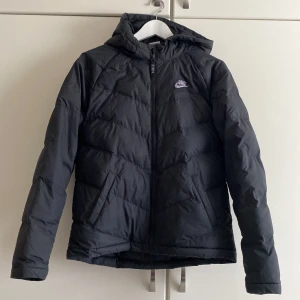 Svart dunjacka med huva från Nike - Säljer en svart dunjacka från Nike med huva och dragkedja framtill. Jackan har diskret Nike-logga på bröstet och snygga sömmar som ger en sportig look. Perfekt för kyliga dagar när du vill hålla dig varm och bekväm.