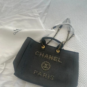 Svart handväska  - Säljer en svart handväska från  med guldiga detaljer och kedjehandtag. Väskan har ett flätat mönster och texten framtill. Invändigt finns dragkedjefack och väskan kommer med dustbag. Perfekt inför stranden