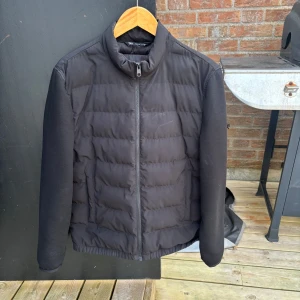 Svart pufferjacka med dragkedja - Säljer en svart pufferjacka från Zara Jackan har hög krage, hel dragkedja framtill och två sidofickor. Ärmarna är släta medan kroppen är quiltad för extra värme. Perfekt för kyliga dagar.