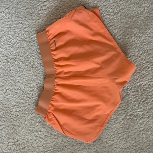 Orange träningsshorts från Adidas - Snygga orange träningsshorts från Adidas med bred resår i midjan och logga på sidan. Lätta och luftiga med sportig design, perfekta för aktiva dagar. Gjorda i syntetmaterial för maximal komfort.