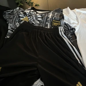 Arsenal svartvit fotbollströja från Adidas - Säljer en svartvit Arsenal fotbollströja från Adidas med unikt grafiskt mönster och guldfärgade detaljer. Tröjan har korta ärmar och klubbens logga på bröstet. Perfekt för dig som vill sticka ut på planen eller på läktaren.