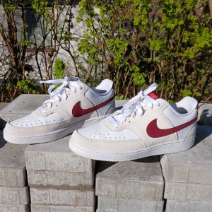 Nike sneakers med röd swoosh - Säljer ett par vita Nike sneakers med röd swoosh och detaljer. Skorna har klassisk design med perforerad tå, vita snören och lågt skaft. Perfekta för dig som gillar stilrena och sportiga skor med en färgklick.