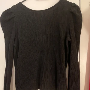 Svart långärmad blus med knyt - Säljer en svart långärmad topp med rynkad struktur och knytning bak. Toppen har lätt puffade axlar. Perfekt att styla med jeans eller kjol.