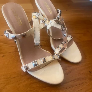 Vita klackskor med nitar - Säljer ett par beige klackskor med remmar dekorerade med silvriga nitar. Säljer de billigt eftersom ena spännet på ena skon är av. 
