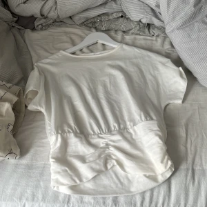 Vit kortärmad topp med rynkdetalj - Säljer en vit kortärmad topp med rund hals och rynkdetalj i midjan som ger en snygg form. Toppen har en enkel och stilren design, perfekt att matcha med jeans eller kjol. Den är helt ny använd ca 2 gånger. Den är inte tvättad men tvättar alltid innan postning. Köp gärna!! Det är storlek 158/164 