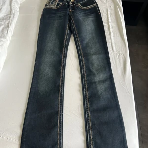 Mörkblå jeansbyxor från True Religion - Säljer ett par mörkblå jeansbyxor från True Religion med tydliga kontrastsömmar och ikoniska bakfickor. Byxorna har bootcut-modell och klassisk femficksdesign. Snygga detaljer med broderade sömmar och logopatch i läder baktill.     Storlek 26.   Aldrig använda bara testade 