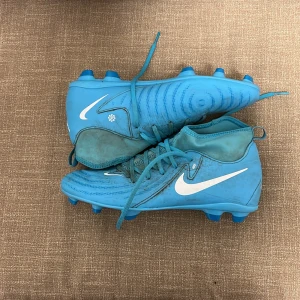 Blå Nike fotbollsskor med snörning - Säljer ett par blå Nike fotbollsskor med vit swoosh-logga på sidan. Skorna har snörning och rund tå, samt dobbar för bra grepp på planen. Perfekta för dig som vill sticka ut på fotbollsplanen!