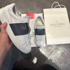 Valentino open  - Säljer ett par vita Valentino Open sneakers med bred svart rem över mitten. Klassisk låg modell med rund tå, vita skosnören och diskret Valentino-logga på plösen. Snygg och stilren design som passar till det mesta. Skulle säga att skicket är 8,5/10. Storlek 41 men de passar 41-42  Perfekt till sommern!!