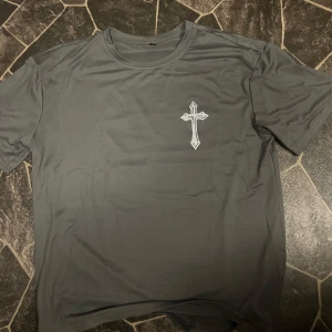 Mörkgrå t-shirt med kors och text - Säljer en mörkgrå t-shirt med ett vitt kors på bröstet och stor röd/vit text på ryggen: 'Believe in what you pray for'. T-shirten har rund hals och korta ärmar. Perfekt för dig som gillar statement-plagg.