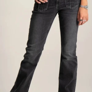 Gråa/svarta bootcut jeans med fickdetaljer - Säljer ett par svarta/gråa low waist bootcut jeans med snygga fickor framtill som har lock och knappar. Använda ett par gånger och är lite lite slitna ner till men inget som märks. Köpta från Gina Young för ca 400 och säljs inte längre.